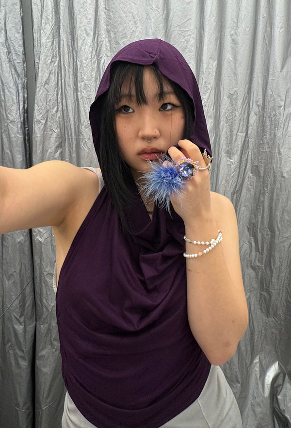 Close-up of a plum purple hooded top with draped neckline. Highlights include a bold blue feathered statement ring and delicate pearl jewelry.