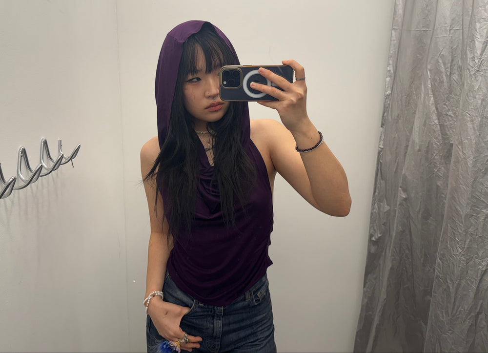 Person wearing a deep purple cowl neck hooded top with a sleeveless halter silhouette. Accessorized with layered bracelets and rings for a modern Y2K-inspired look.