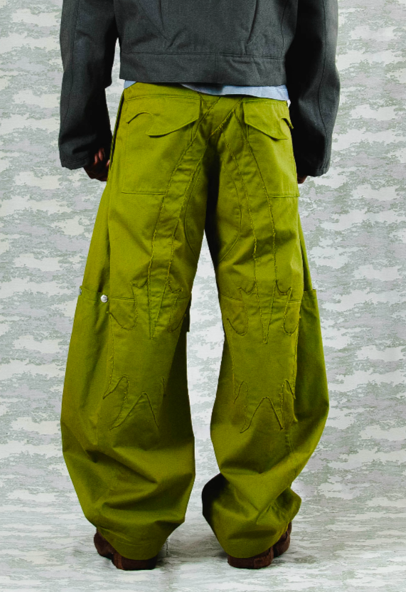Back view of oversized olive green cargo pants with abstract stitching and wide legs – styled with a cropped dark grey jacket, against a camouflage-patterned background