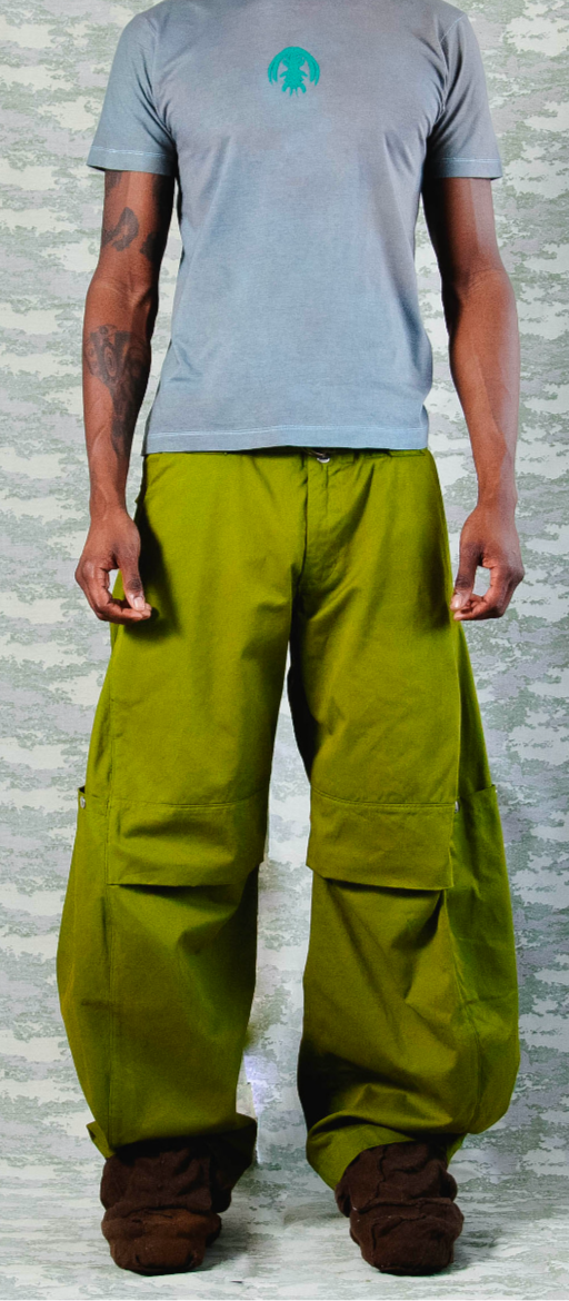 Front view of model wearing wide-leg olive green cargo pants with utility pockets, paired with a fitted grey t-shirt featuring a turquoise chest graphic – set against a camo-inspired backdrop