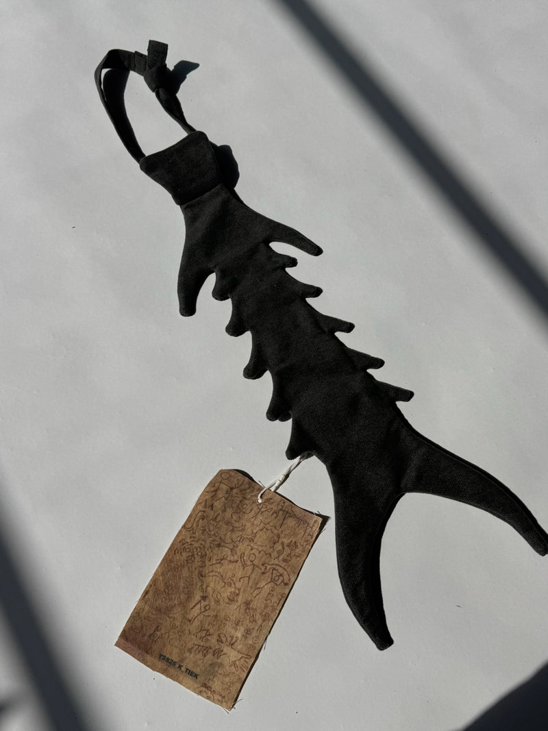 Flat lay of dark grey sculptural fabric accessory shaped like a fish skeleton, featuring soft textile ribs and wide forked tail with illustrated brown paper tag attached – conceptual handmade piece by YAKU