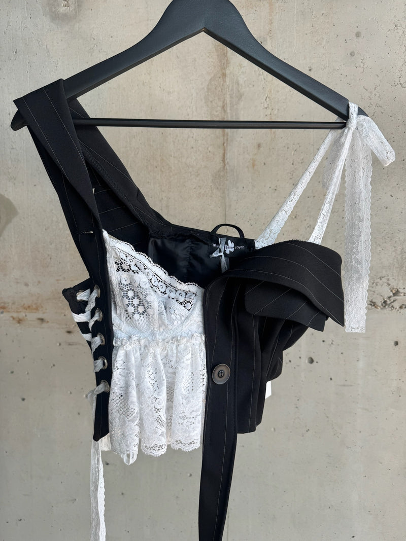 Detailed front view of an avant-garde asymmetrical corset top by Gloria Jane Royer, blending black pinstripe suiting elements with white lace and ribbon straps, styled on a hanger against a concrete wall