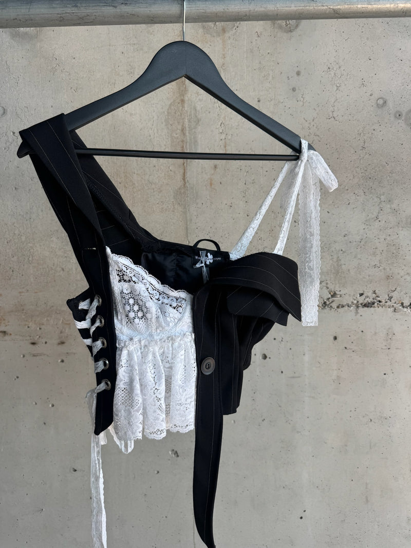 Side view of a deconstructed asymmetrical top by Gloria Jane Royer, combining black pinstripe suiting fabric with intricate white lace and corset-style lacing, hanging against a raw concrete wall