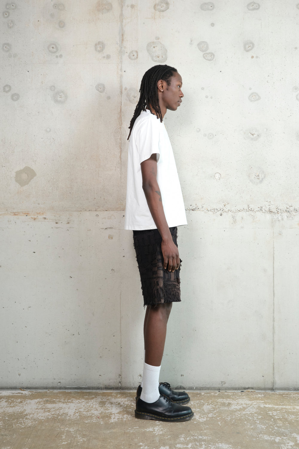 Model in a relaxed pose, wearing a minimalist outfit featuring a white oversized T-shirt, black textured shorts with a deconstructed design, white socks, and black leather shoes. Accessories include a gold pendant necklace and rings. The model stands against a raw concrete wall.