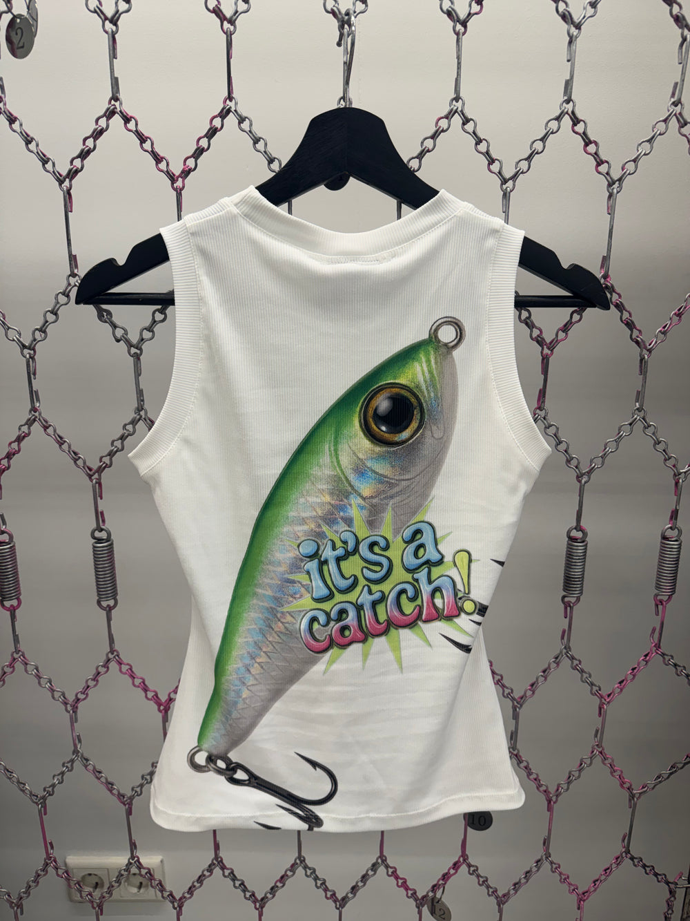White sleeveless shirt with a fishing lure design and 'it's a catch!' text on a patterned background.