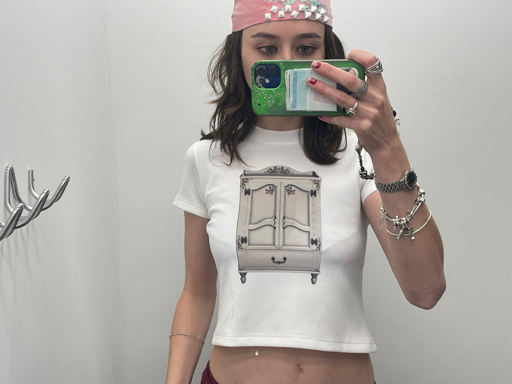 Person wearing a white t-shirt with a graphic design, holding a phone, against a plain background.