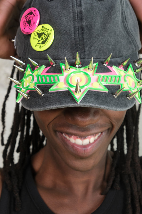 Close-up of a smiling model wearing a black cap adorned with neon green and pink spiked 3D star embellishments and colorful graphic pins.