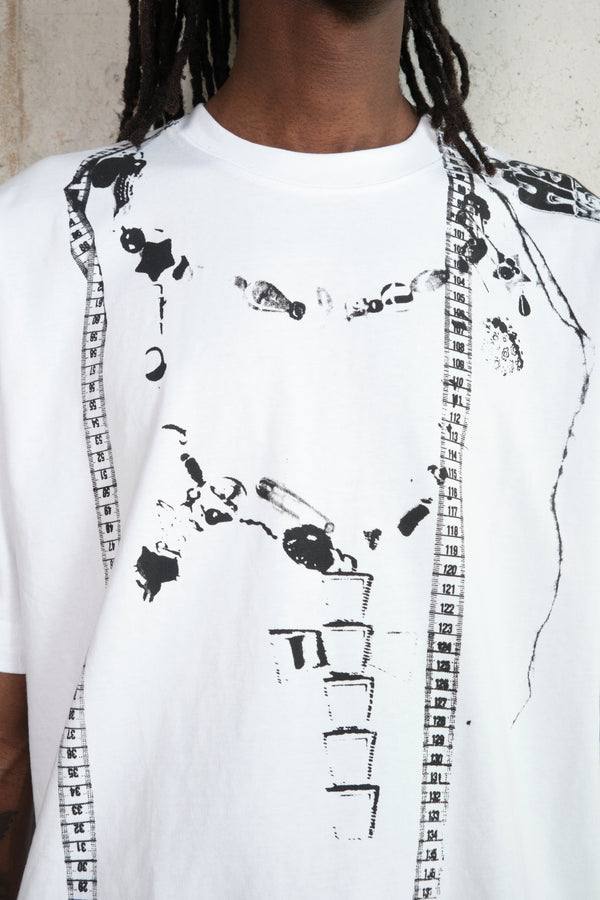 Close-up of a white T-shirt featuring a black abstract print of measuring tapes, jewelry, and scattered objects, worn by a person with dark skin and long dreadlocks, photographed against a concrete wall background.