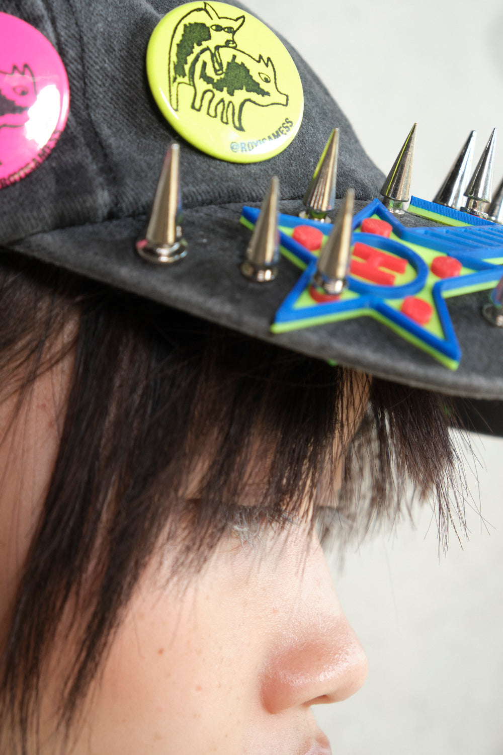 Close-up side view of a person wearing a black cap with neon yellow pin reading “@ROYISAMESS”, colorful 3D star embellishment, and silver spikes, highlighting the eye and fringe against a concrete wall background.