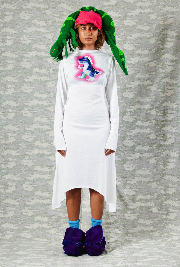 Model in long-sleeve white asymmetrical dress with neon dinosaur print, styled with a bold pink and green sculptural headpiece and chunky purple shoes – playful avant-garde look against camo-patterned backdrop
