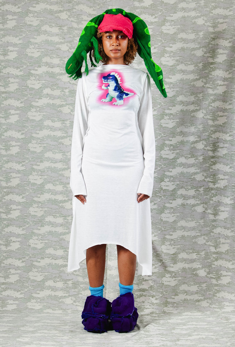 Model in long-sleeve white asymmetrical dress with neon dinosaur print, styled with a bold pink and green sculptural headpiece and chunky purple shoes – playful avant-garde look against camo-patterned backdrop