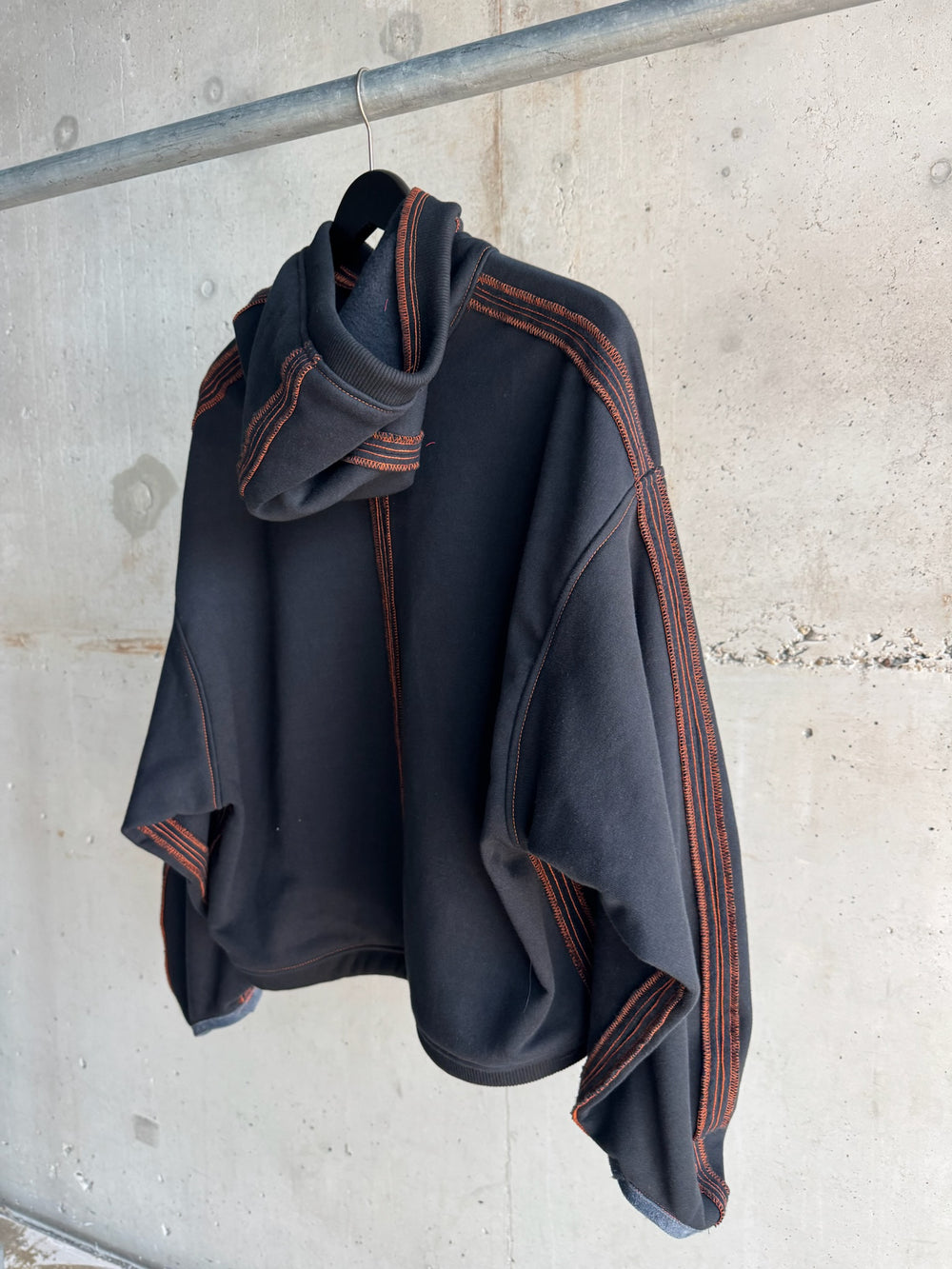 Back view of a black cropped hoodie with bold orange contrast stitching along the seams and hood, displayed on a hanger against a raw concrete wall backdrop