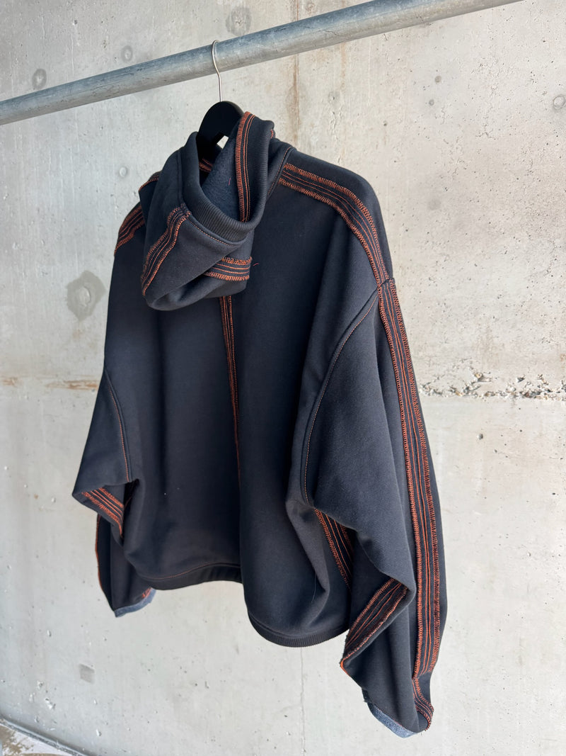 Back view of a black cropped hoodie with bold orange contrast stitching along the seams and hood, displayed on a hanger against a raw concrete wall backdrop