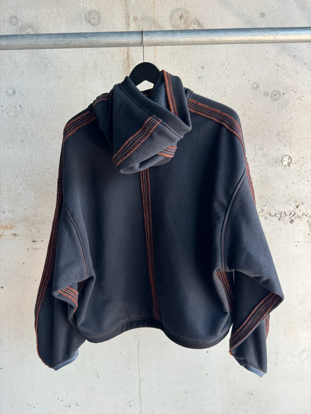 Back view of a black hoodie with oversized sleeves, bold orange contrast stitching along seams and hood, and a relaxed cropped fit, displayed on a hanger against a concrete wall background
