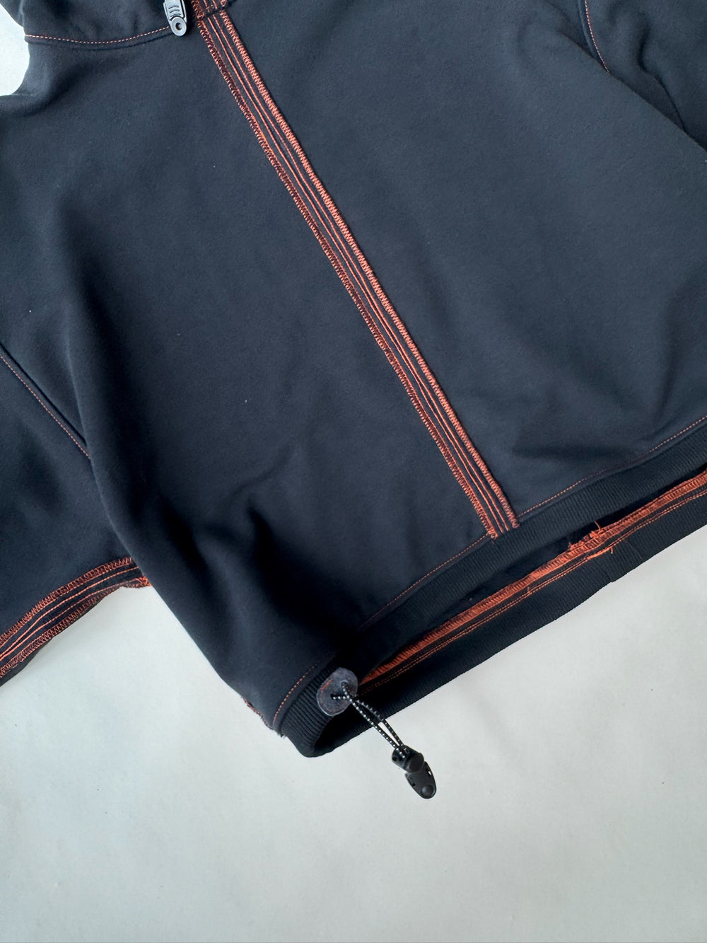 Close-up of the hem and zipper detail on a black cropped hoodie with orange contrast stitching and an adjustable drawstring toggle, displayed on a light background