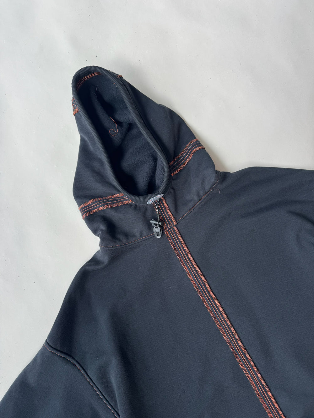 Close-up of the hood and upper chest of a black cropped hoodie with orange contrast stitching and a half-zip front, displayed on a light surface
