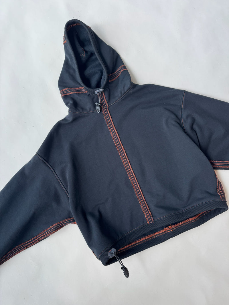 Flat lay of a black cropped hoodie with a front zip and hood, featuring exposed orange contrast stitching and an adjustable drawstring hem, displayed on a light neutral background