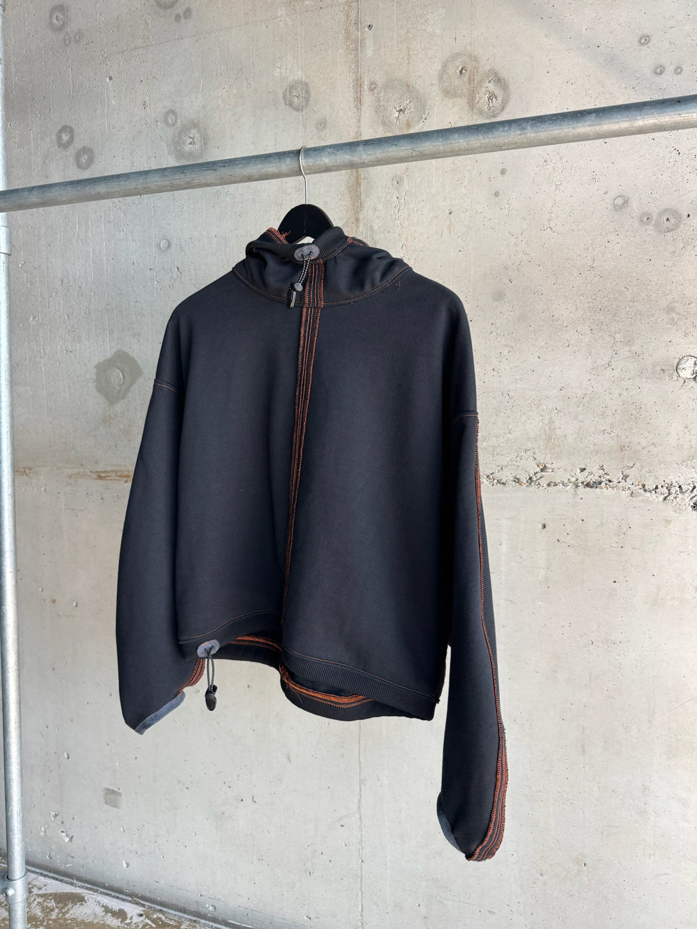  Front view of a black cropped hoodie with front zip, drawstring hem, and bold orange contrast stitching, hanging on a black hanger against an industrial concrete wall