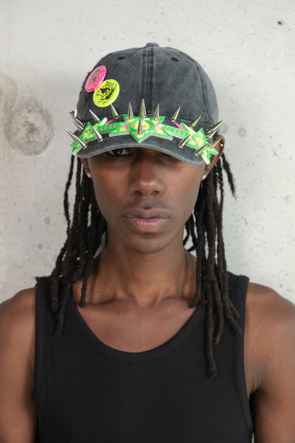 Model wearing a black tank top and a distressed black cap adorned with neon green 3D star spikes and colorful graphic pins, standing against a concrete wall.