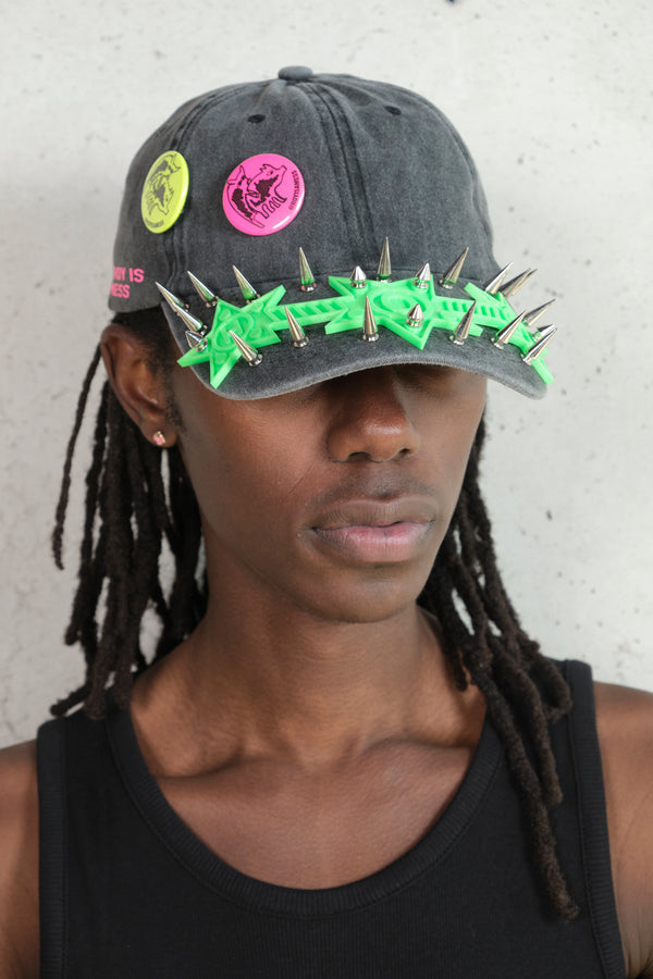 Close-up of model wearing a distressed black cap with neon green plastic arrows, silver spikes, and colorful graphic pins reading ‘Everybody Is Priceless,’ styled with a black tank top.