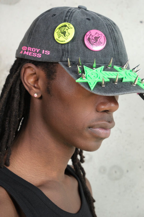 Side profile of model wearing a distressed black cap with neon green plastic embellishment and silver spikes, featuring colorful graphic pins and pink text ‘@ROY IS A MESS,’ styled with a black tank top