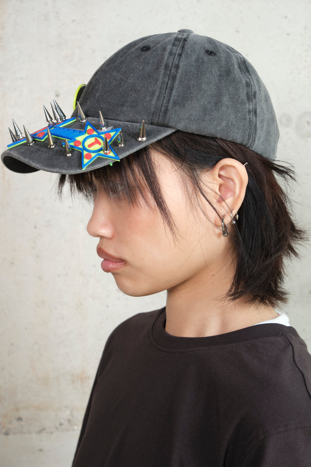 Side profile of a person wearing a black cap with colorful 3D star motifs and silver spikes, paired with layered silver earrings and a dark oversized t-shirt, set against a concrete wall background.
