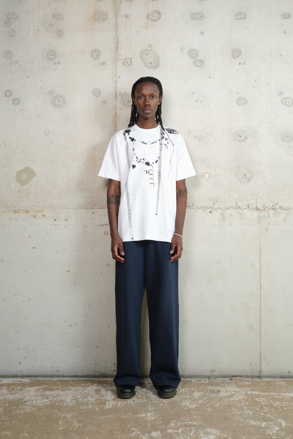 Model standing against a raw concrete wall, wearing a white oversized graphic T-shirt with black abstract prints and measuring tape details, paired with wide-leg navy trousers and black shoes, showcasing a minimal yet artistic streetwear look