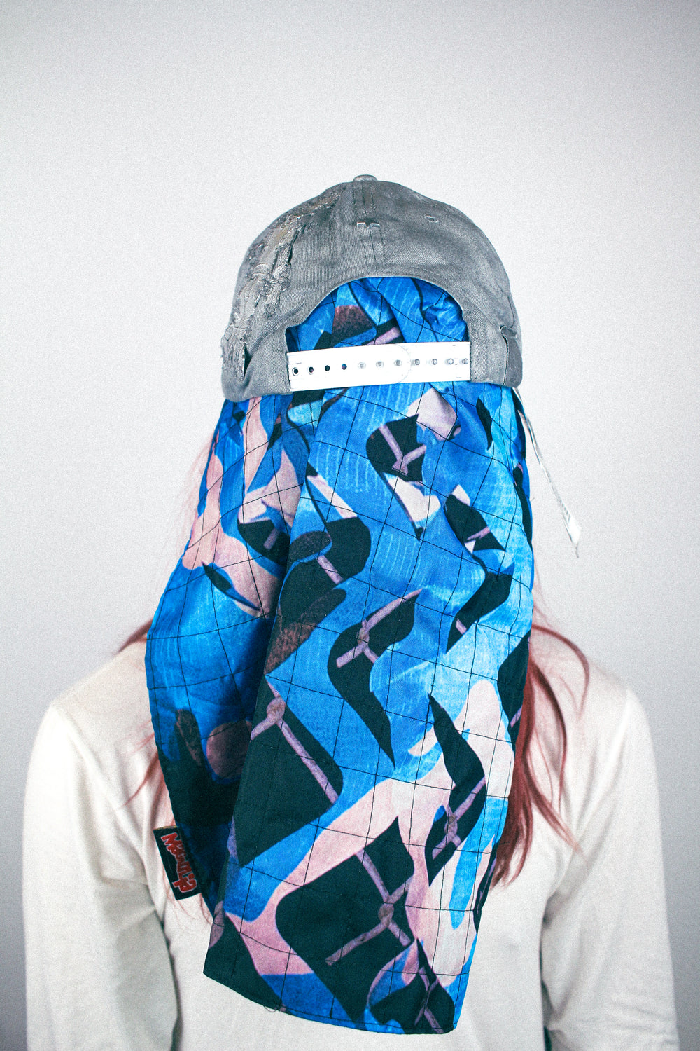 Grey distressed baseball cap worn with quilted scarf panel draped at the back in blue and pink graphic print.
