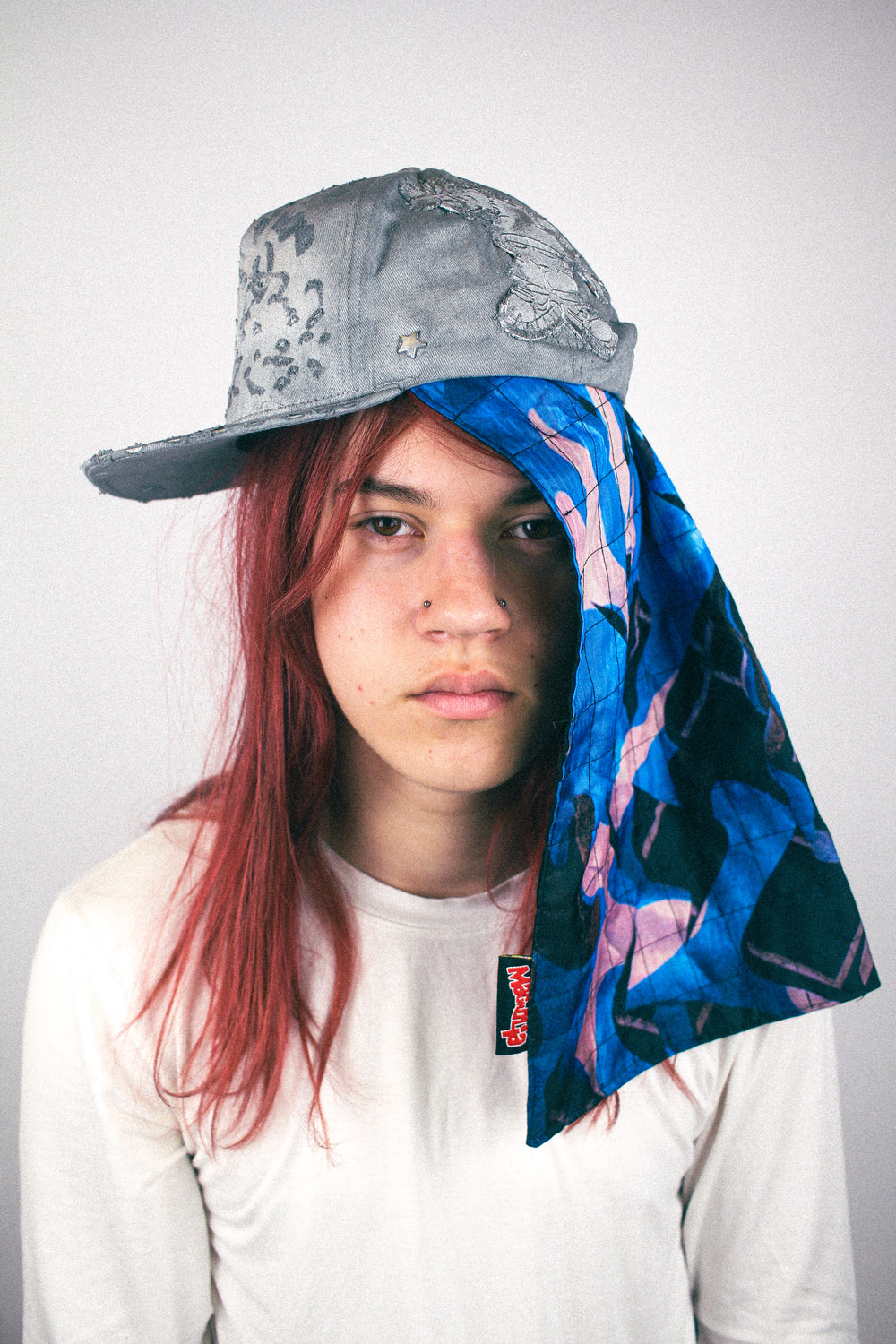 Grey distressed baseball cap worn with quilted scarf panel draped at the side in blue and pink print.