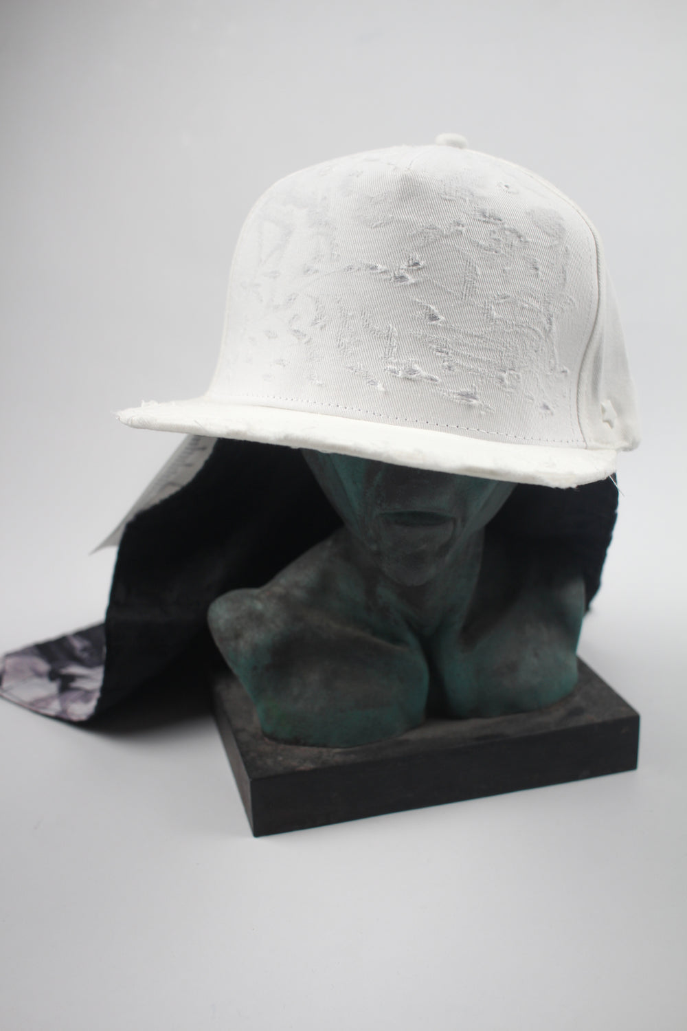 White distressed baseball cap with flat brim and worn fabric texture, displayed on sculptural stand.