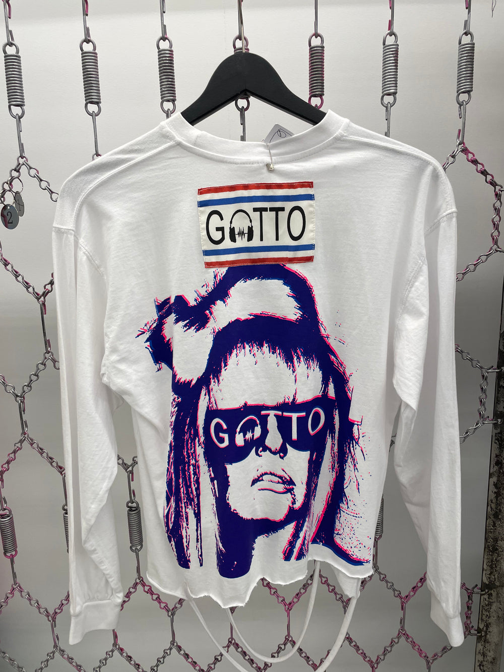 White shirt with graphic design and 'GOTTO' branding on a hanger against a textured wall.