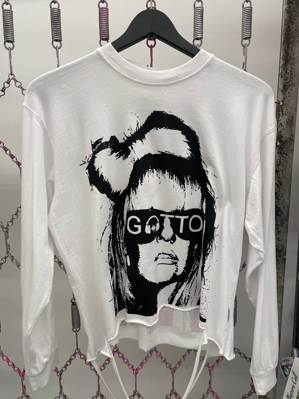 White sweatshirt with black graphic design and 'Gatto' text on a hanger against a chain-link fence background.