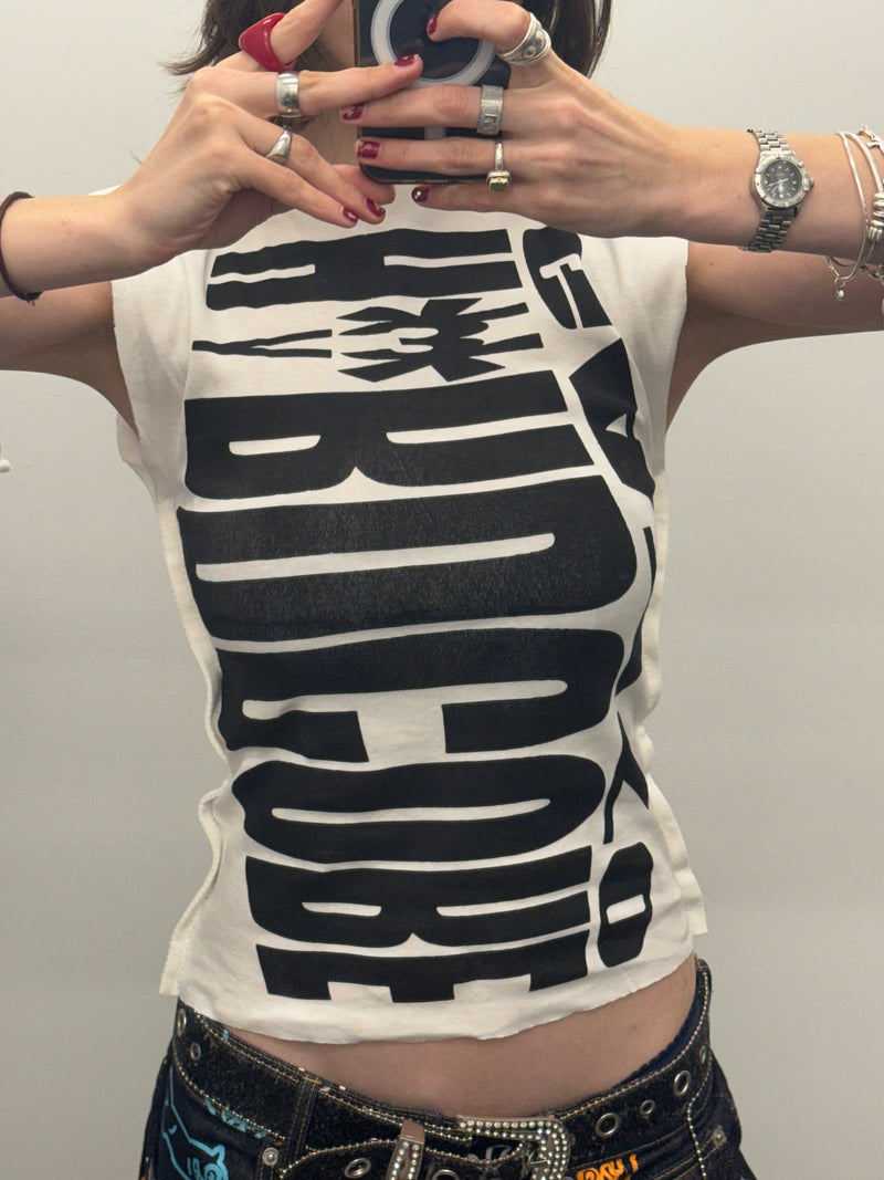 Person wearing a white sleeveless shirt with black text design, holding phone with both hands.