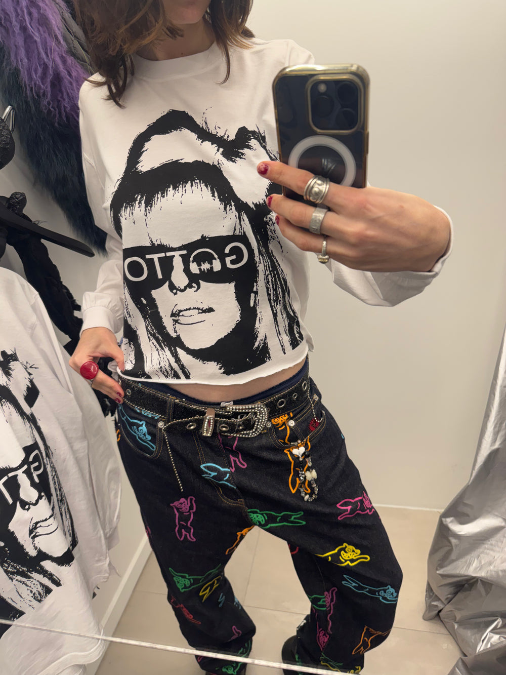 Person wearing a white t-shirt with a graphic design and colorful pants, taking a mirror selfie.