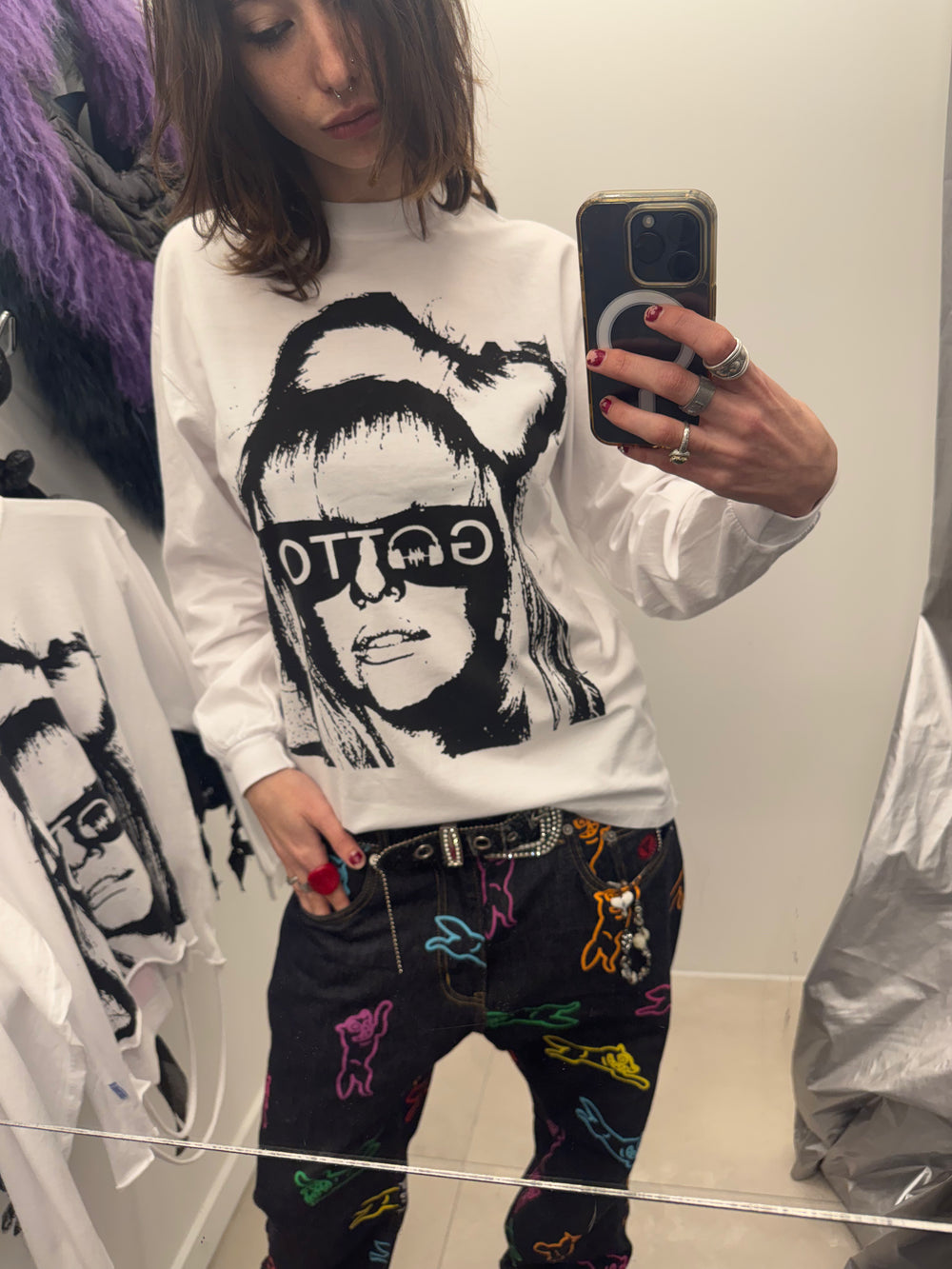 Person wearing a white sweatshirt with a graphic design and colorful pants, taking a mirror selfie.