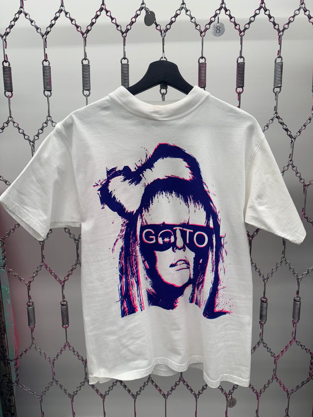 White t-shirt with a graphic design of a person with glasses and text, hanging on a chain link fence.