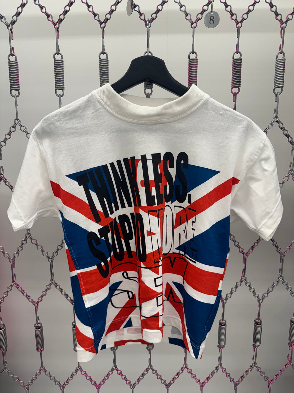 White t-shirt with Union Jack design and text on a hanger against a chain-link fence background