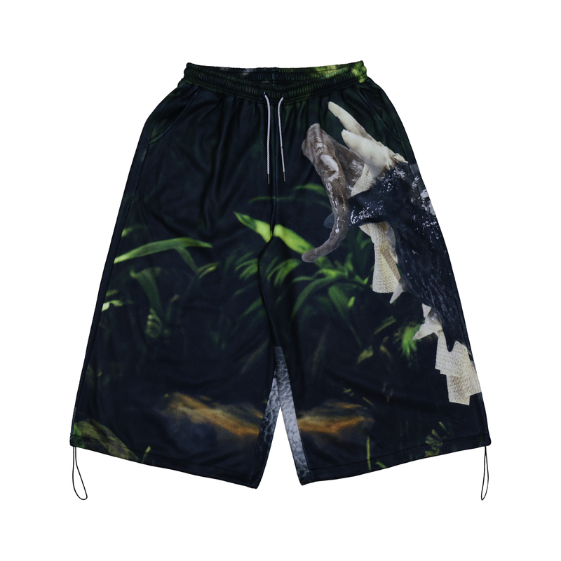 Long shorts featuring a bold nature-inspired print, depicting lush green foliage and a photorealistic image of an animal’s open mouth. The design has a dark, earthy tone with drawstrings on the waistband and hems, creating an adjustable, relaxed fit that merges outdoor aesthetics with urban style.