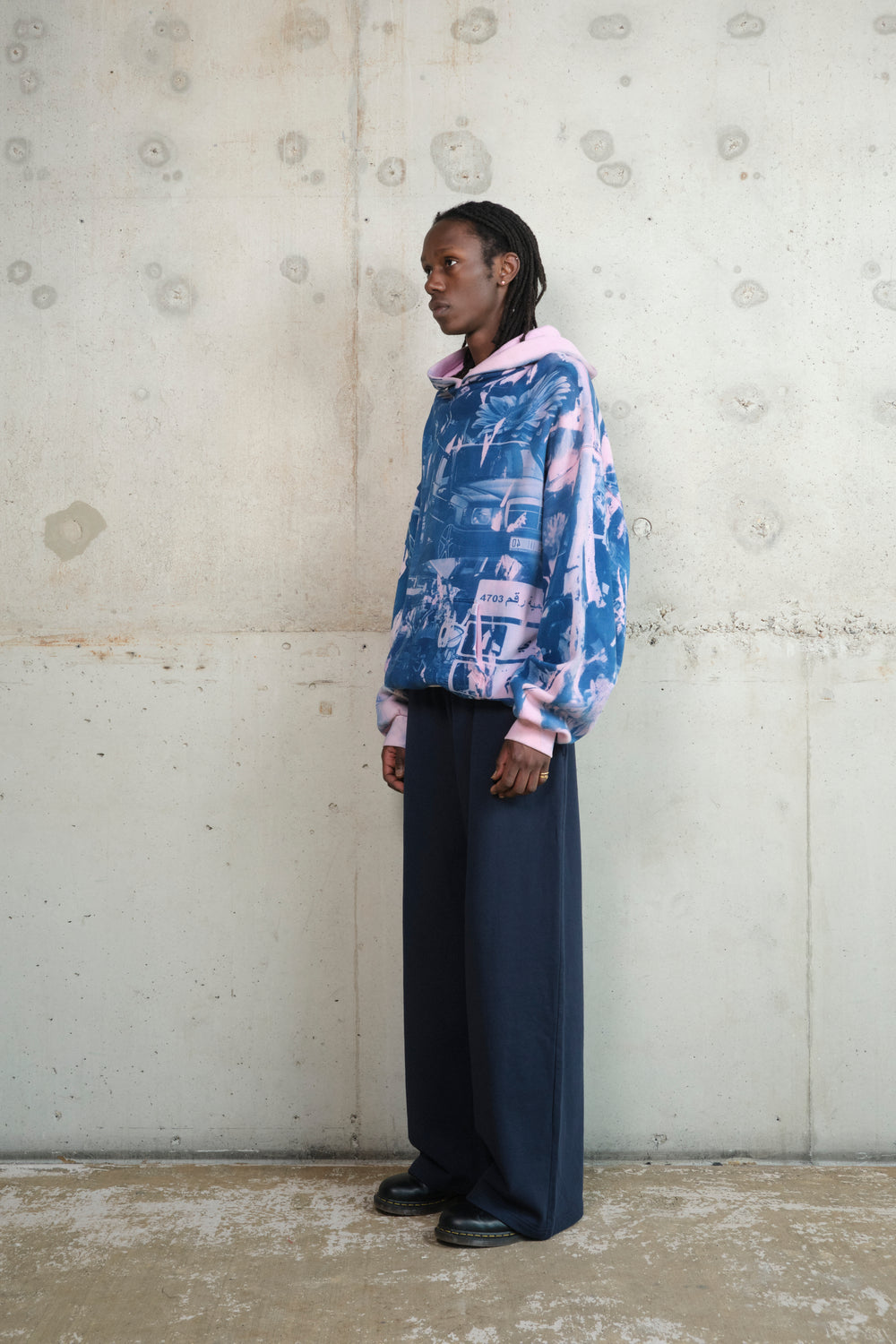 Side profile of model wearing oversized pink and blue graphic hoodie with layered collage print, styled with high-waisted wide-leg navy pants and black shoes – minimalist concrete backdrop