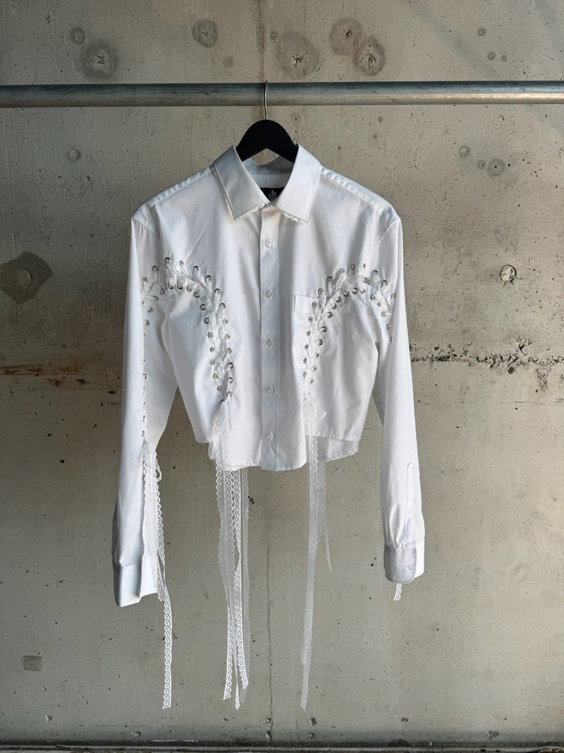 White cropped button-up shirt with silver eyelet lace-up detailing and long decorative white ribbons, displayed against a concrete wall on a hange