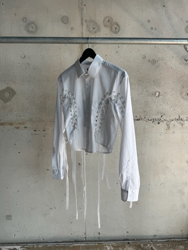 White long-sleeve cropped shirt with lace-up corset detailing and decorative lace ribbons, displayed on a hanger against a raw concrete wall