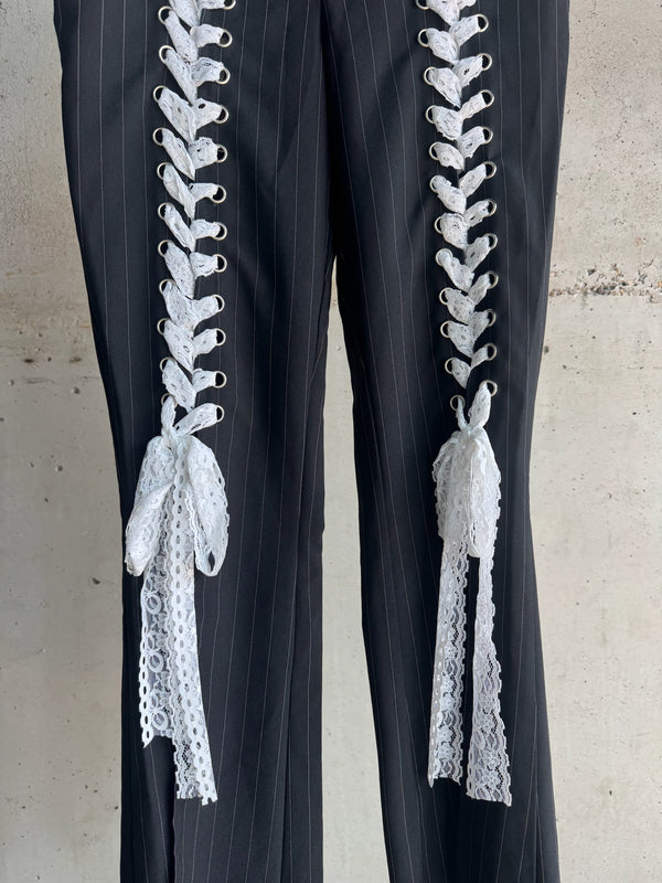 Close-up of black pinstripe trousers featuring intricate white lace-up detailing with decorative bows