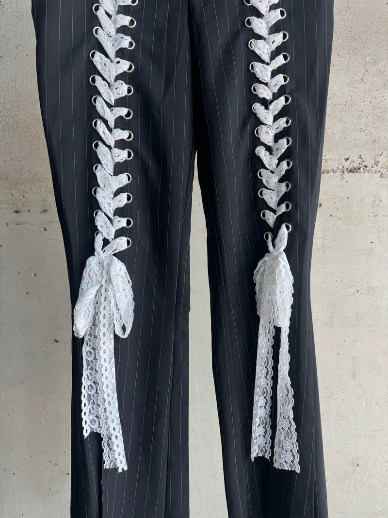 Close-up of black pinstripe trousers featuring intricate white lace-up detailing with decorative bows