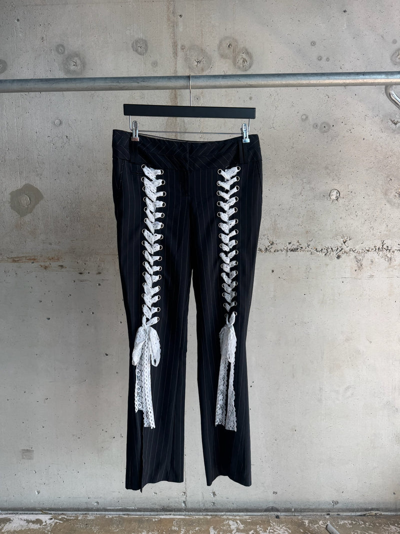 Black pinstripe trousers with white lace-up details and bows displayed on a hanger against a minimalist concrete background