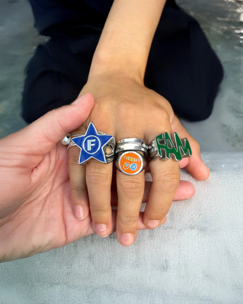 Two hands wearing colorful rings with visible letters on a neutral background