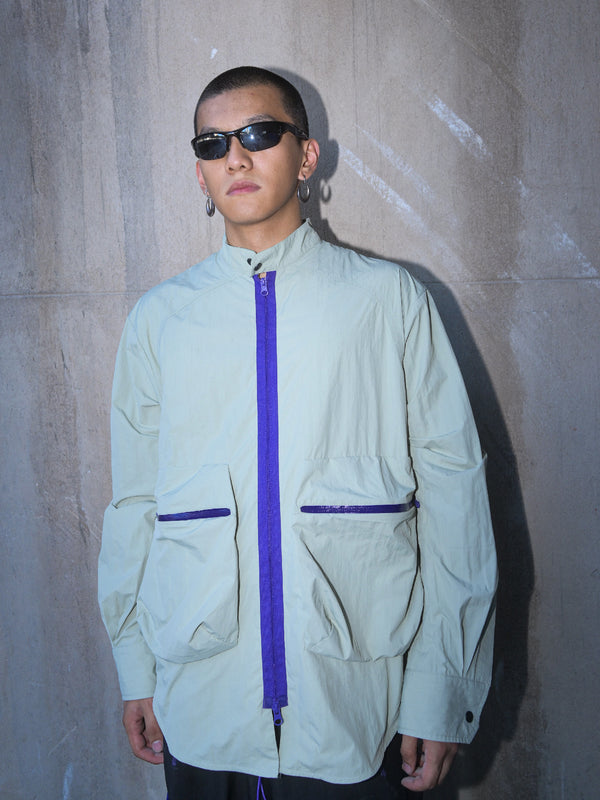 Model in sunglasses against concrete wall
“Model wearing sage green technical overshirt with contrasting purple zipper and oversized utility pockets, styled with black sunglasses and silver hoops – urban outdoor look against raw concrete wall