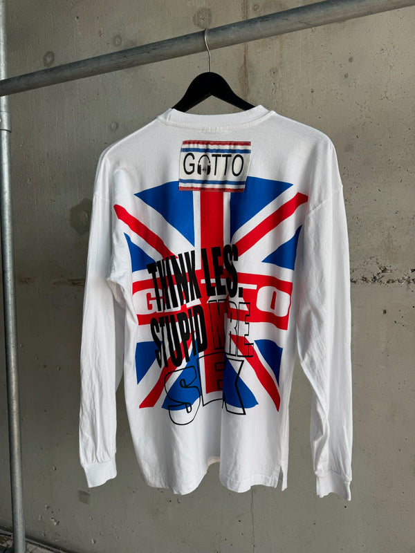 Back view of a white long-sleeve T-shirt with inverted bold text “THINK LESS STUPID MORE SEX” over a Union Jack flag graphic, featuring a stitched “GATTO” label near the collar, hanging on a black hanger against a concrete wall.