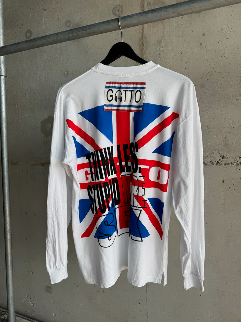 Back view of a white long-sleeve T-shirt with inverted bold text “THINK LESS STUPID MORE SEX” over a Union Jack flag graphic, featuring a stitched “GATTO” label near the collar, hanging on a black hanger against a concrete wall.