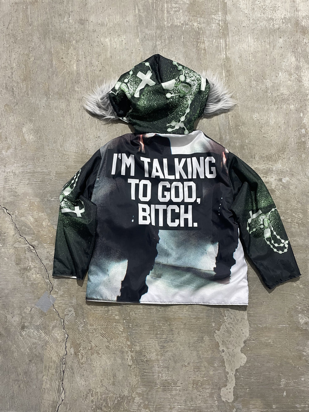 Back view of black hooded padded winter jacket with faux fur trim, featuring bold graphic text print “I’m talking to God, bitch” and green rosary cross artwork on sleeves and hood, statement streetwear jacket.