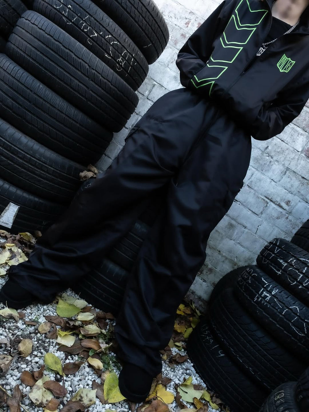 Person wearing a black jacket with green accents standing next to stacked tires on a concrete surface.